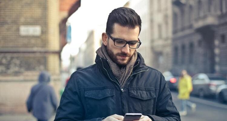 Man Wearing Black Zip Jacket Holding Smartphone Surrounded by Grey Concrete