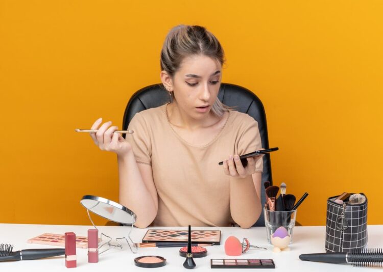 Surprised young beautiful girl sits at table with makeup tools holding makeup brush and looking at phone in her hand isolated on orange background
